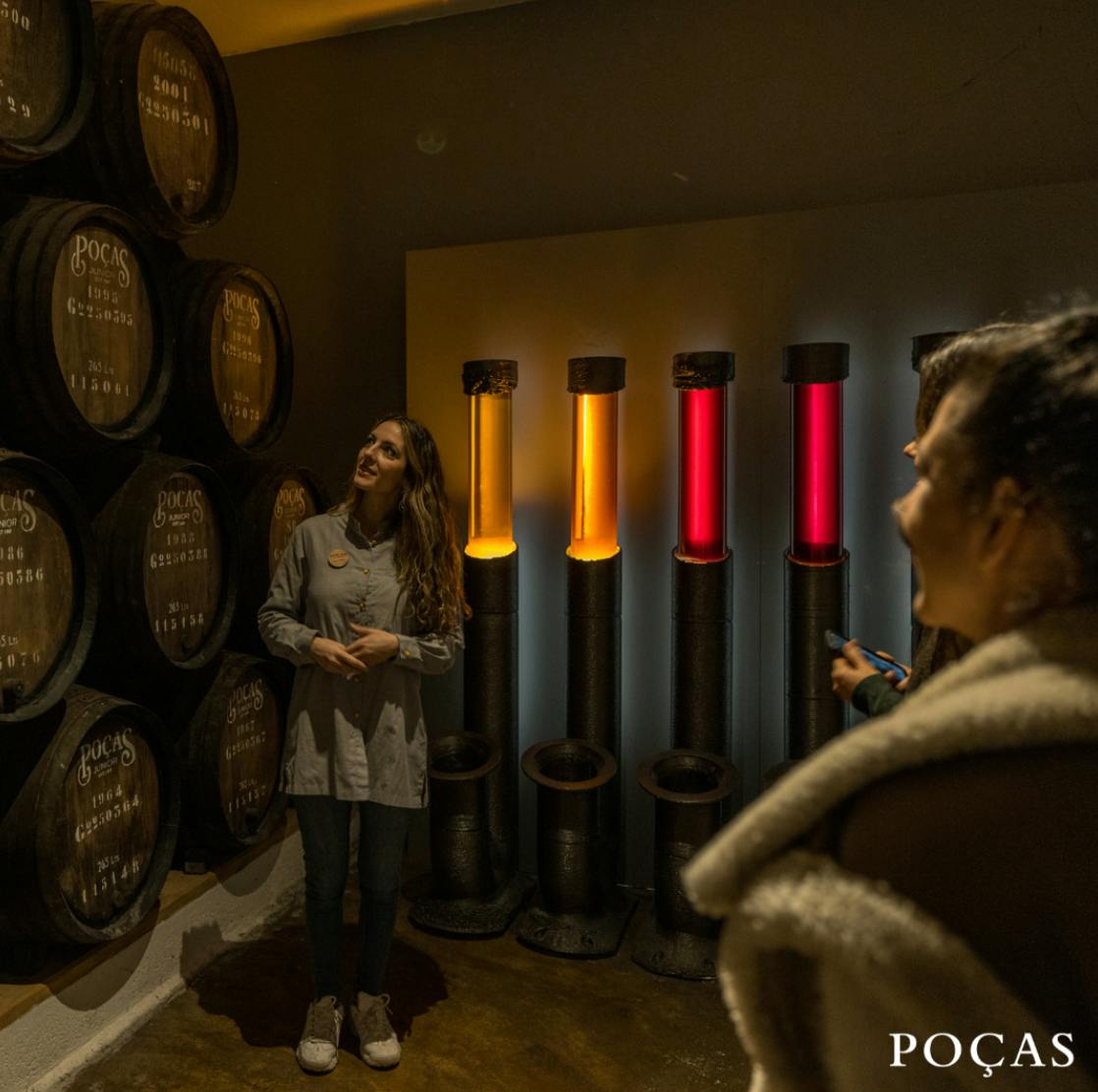 Una mujer de pie cerca de barriles y tubos iluminados, con otras personas observando, en un entorno de bodega.
