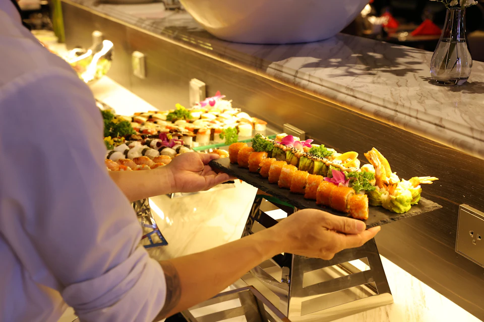 A person arranges a tray of assorted sushi rolls on a buffet table laden with brightly colored sushi and garnishes.
