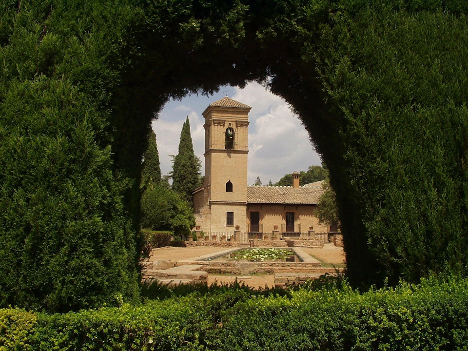 A garden with manicured hedges framing a stone building with a bell tower in the background.