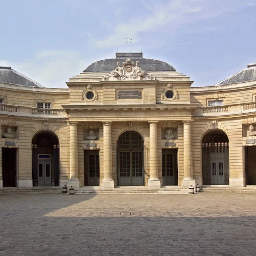 A historic building with a curved facade, arched windows, and columns, set in a cobblestone courtyard under a clear sky.