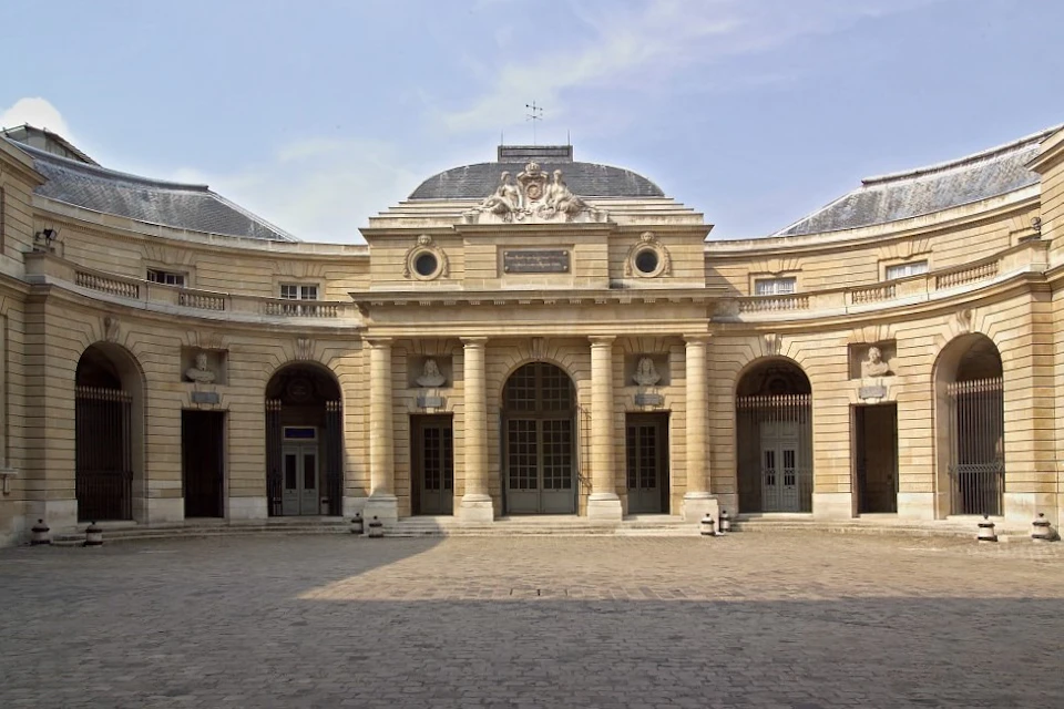 A historic building with a curved facade, arched windows, and columns, set in a cobblestone courtyard under a clear sky.