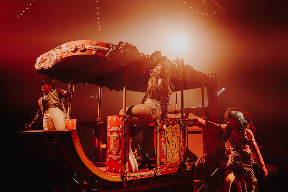 Performer seated on a vibrant, ornate carriage under bright stage lighting, with supporting cast in theatrical costumes.