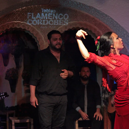 En flamencodansare i en röd klänning uppträder passionerat på scenen, flankerad av musiker mot en bakgrund med "Tablao Flamenco Cordobes" -text.