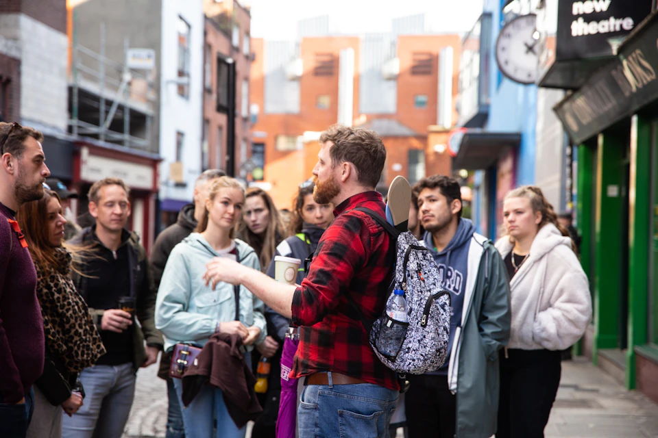 A man in a red plaid shirt talks to a group of people on a city street, holding a cup and wearing a backpack with a skateboard.