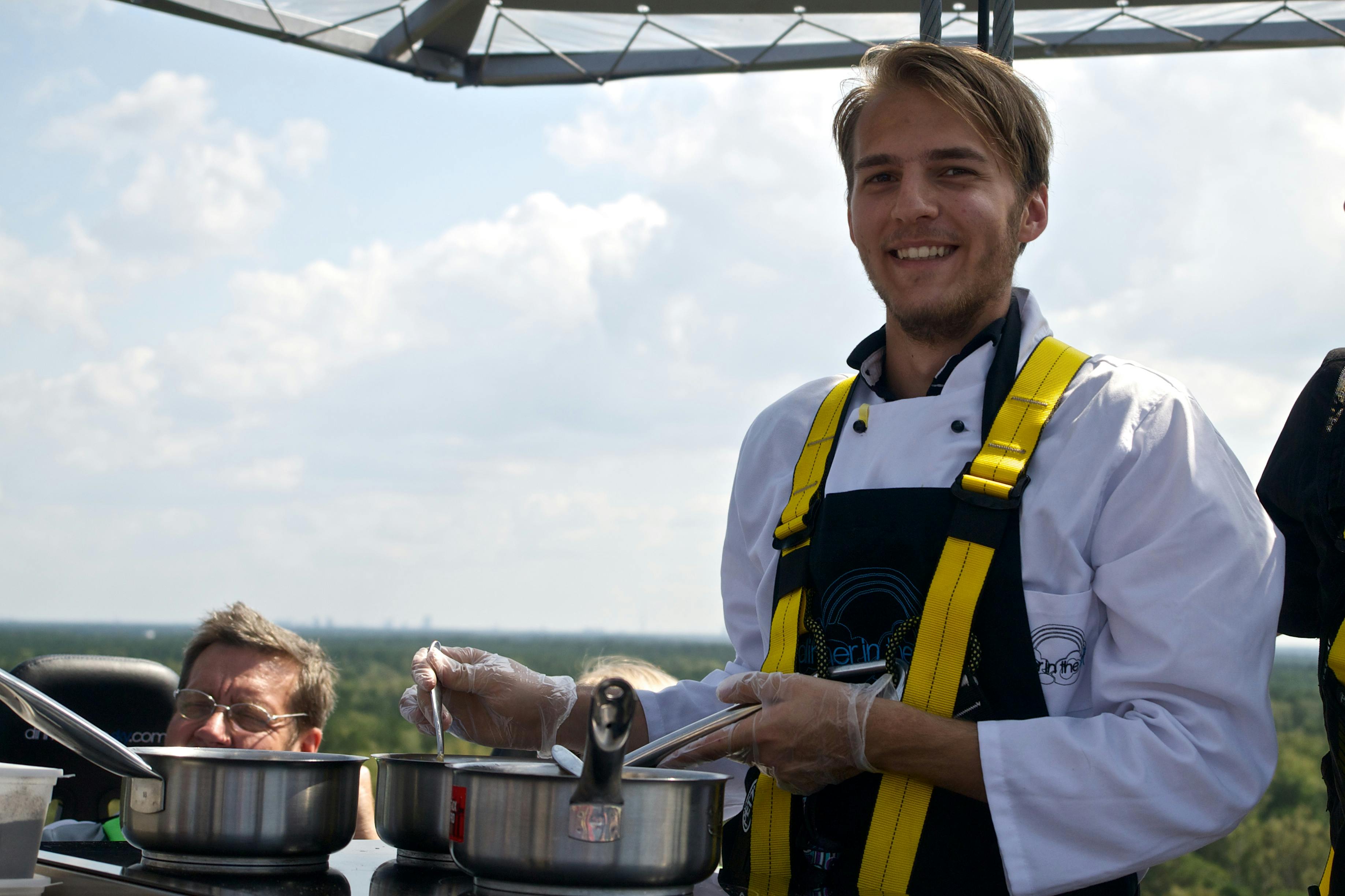 Leende kock i vit rock och harness matlagning i två stekpannor utomhus, med en person som kikar över disken.