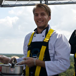 Leende kock i vit rock och harness matlagning i två stekpannor utomhus, med en person som kikar över disken.