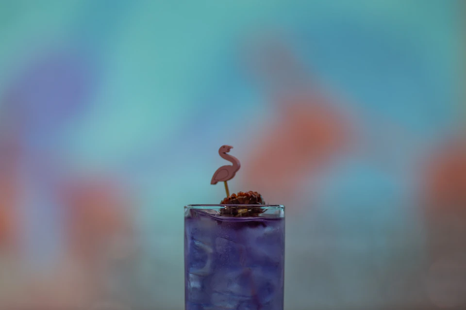 A glass of purple drink with ice and a flamingo-shaped stirrer, set against a blurred colorful background.