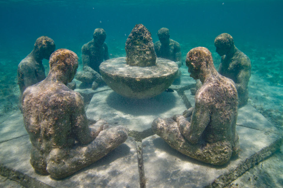Underwater sculptures of seated human figures surrounding a central pedestal in clear blue water.