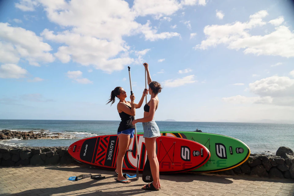 Two people stand on a beach holding paddles, with red and green stand-up paddleboards in the background under a blue sky.
