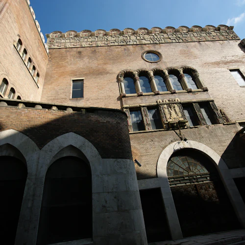 Tegelbyggnad med välvda dörröppningar i sten, prydnadsfönster och ett cirkulärt fönster på övre plan, mot en blå himmel.