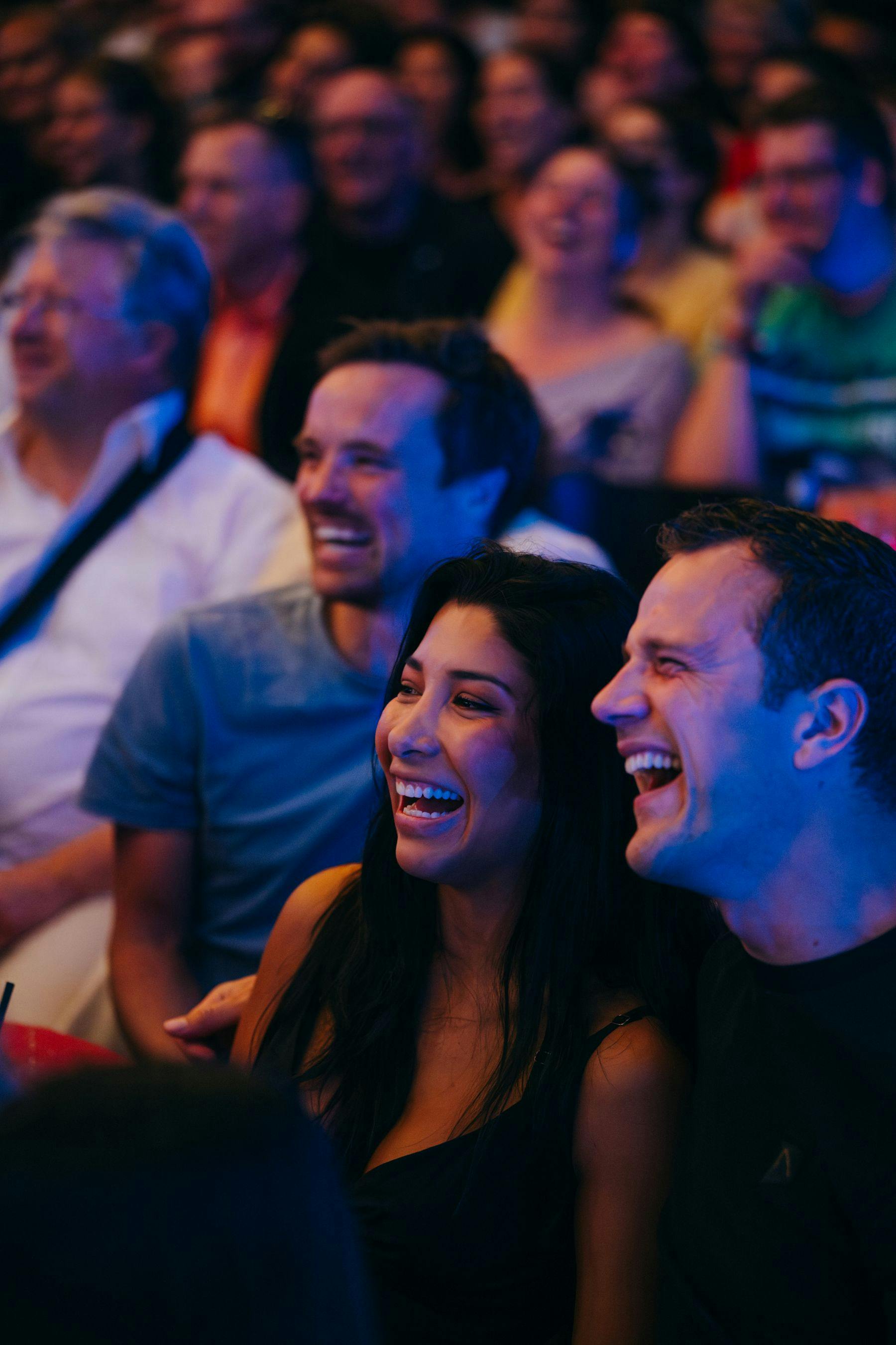 Eine Gruppe von Menschen, die eng beieinander sitzen, lächeln und lachen in einer lebhaften Atmosphäre mit bunter Beleuchtung.