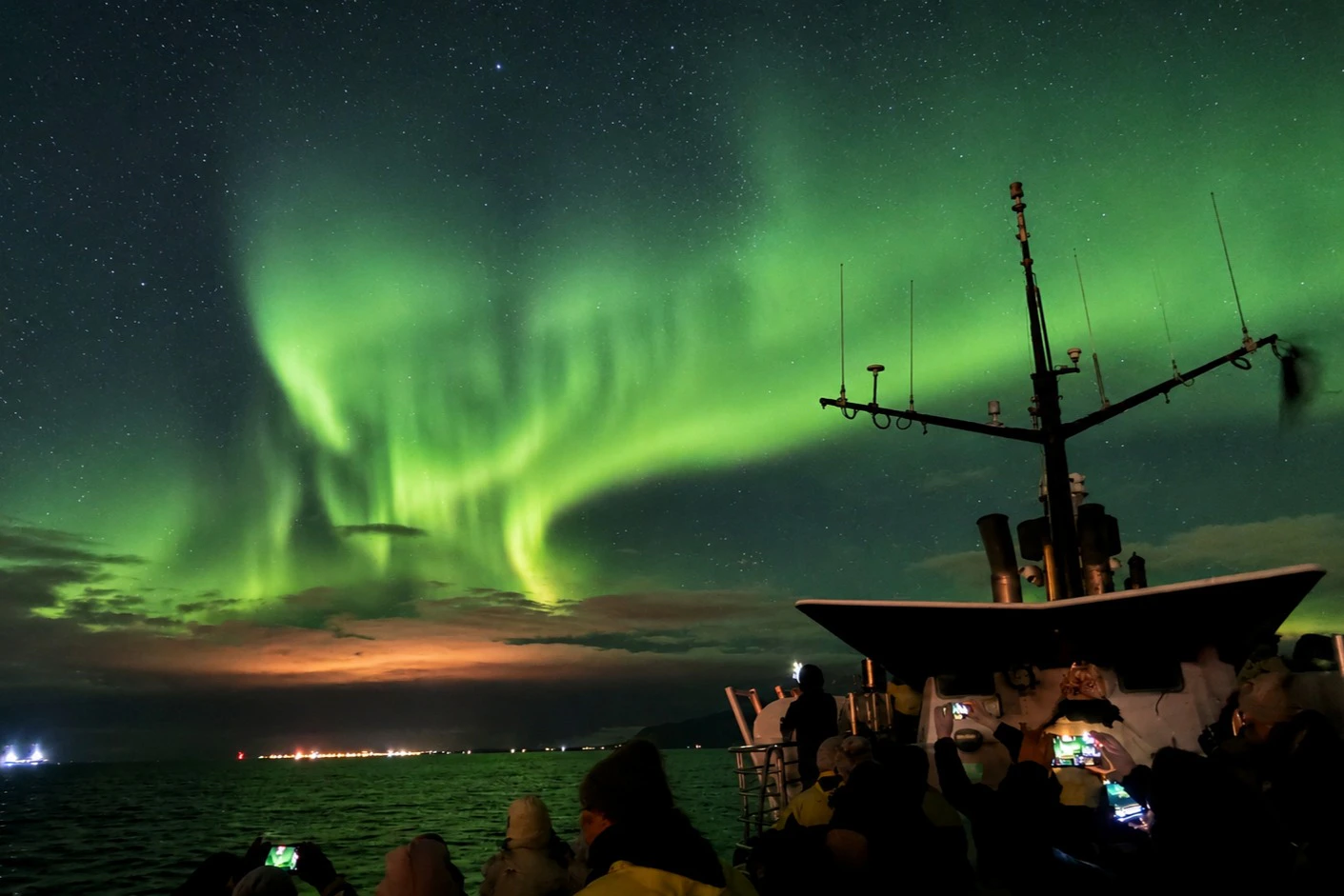 People observing the northern lights from a ship at night, with a green aurora borealis glowing in the sky.