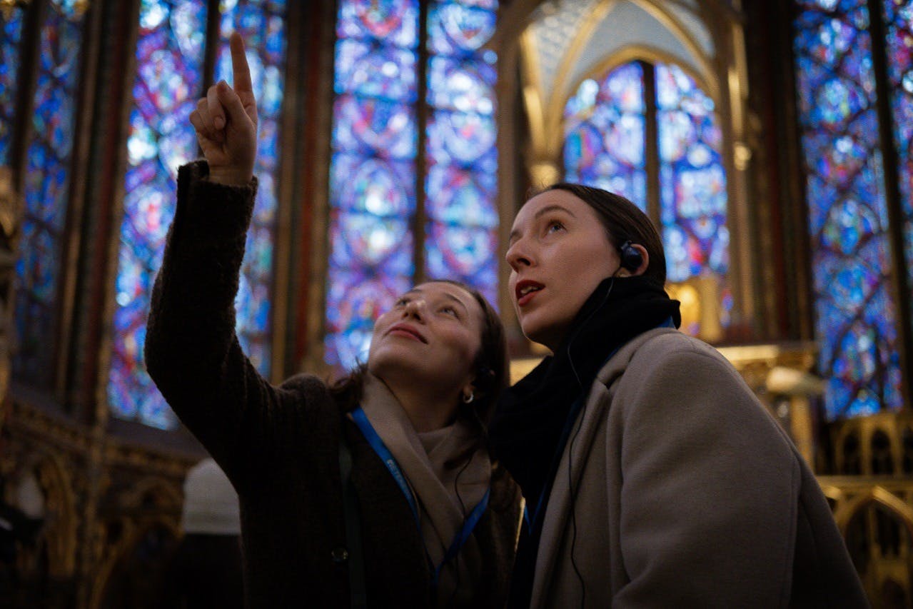 Two women with headsets look upwards in front of colorful stained glass windows inside a cathedral.