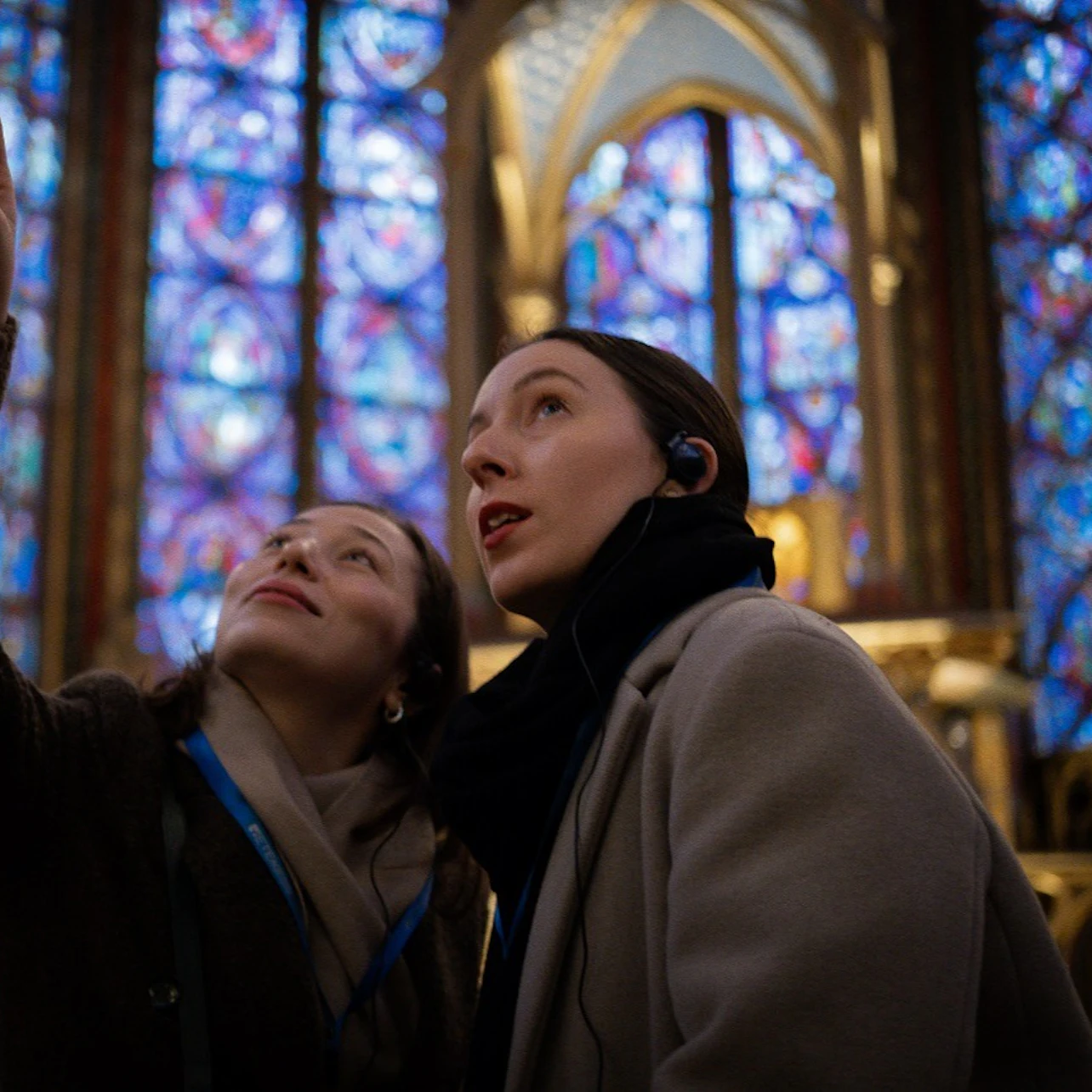 Two women with headsets look upwards in front of colorful stained glass windows inside a cathedral.