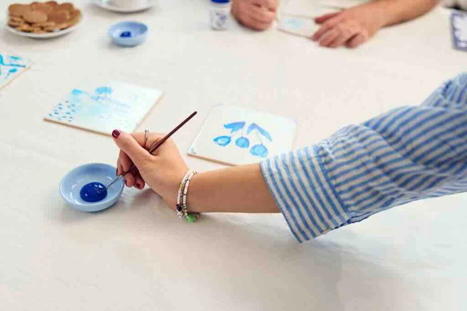 Person in a striped shirt painting blue designs on a tile, with small dishes of blue paint and another person in the background.