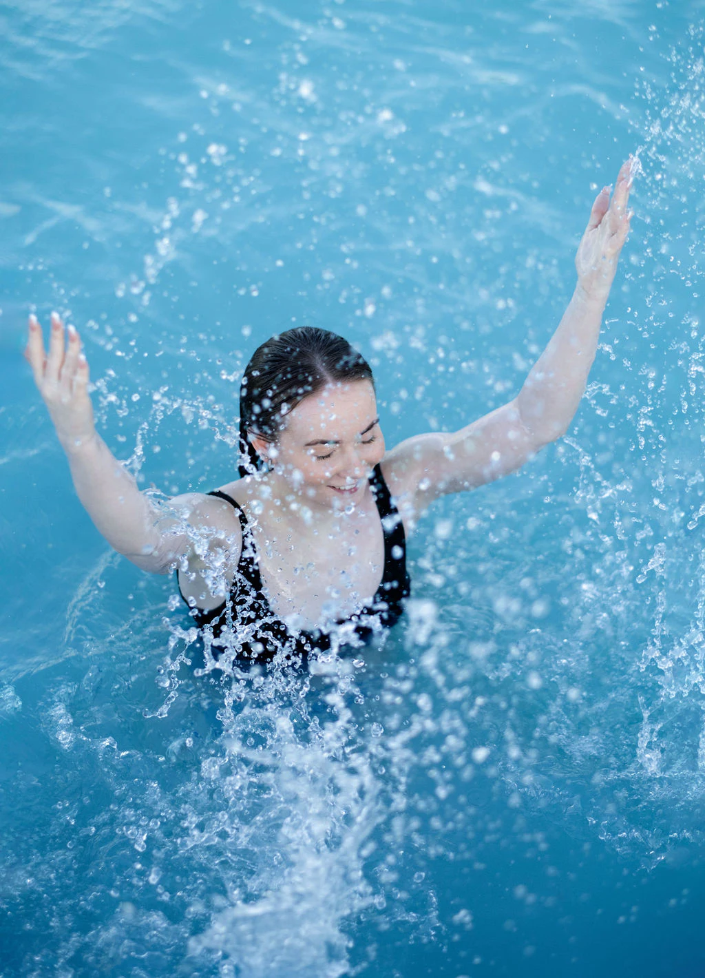 A person in a black swimsuit is joyfully splashing water while standing in a pool with their arms raised.