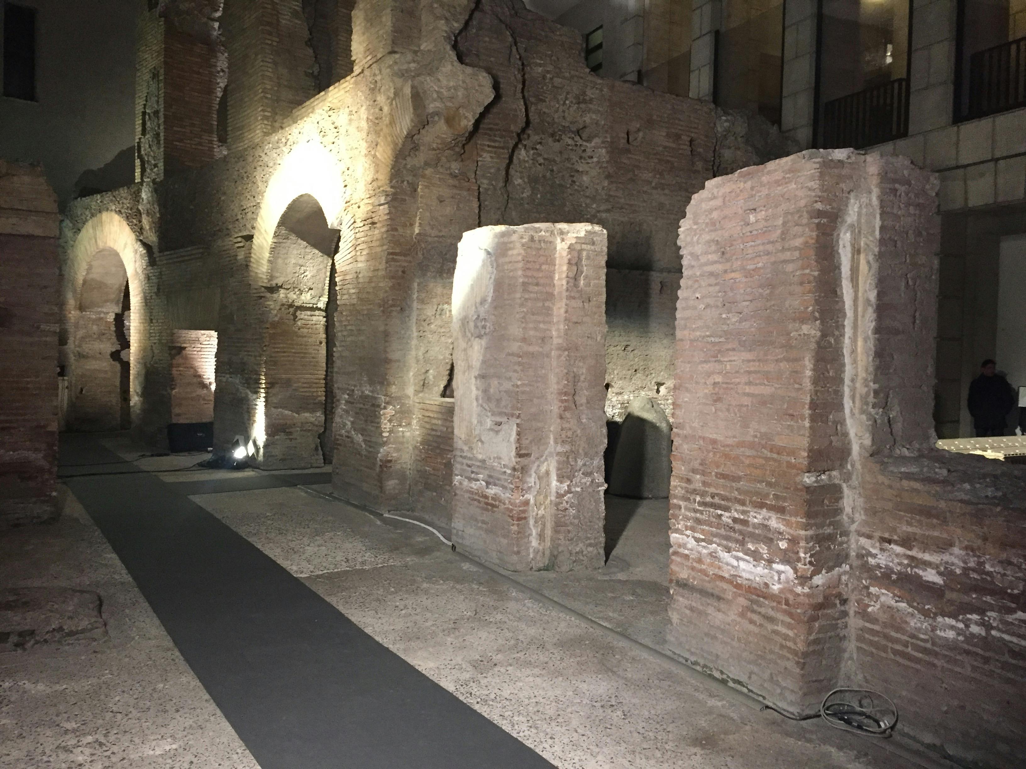 Ancient brick ruins with archways and pillars, illuminated by spotlights at night, surrounded by a concrete floor.