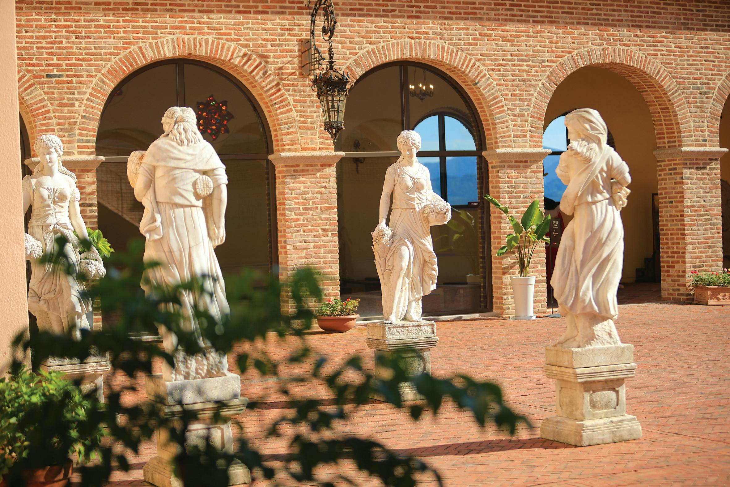 Three white stone statues in a courtyard with brick arches, visible greenery, and a hanging light fixture.