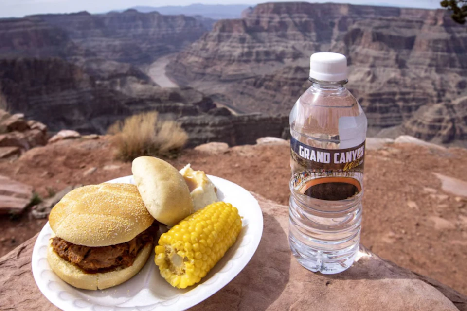 A plate with food, including a sandwich and corn, next to a water bottle labeled 'Grand Canyon,' overlooking a canyon landscape.
