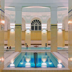 Inomhusspa med gula kakelpelare, en central liten pool och en större pool i bakgrunden. Fyra personer syns i vattnet.