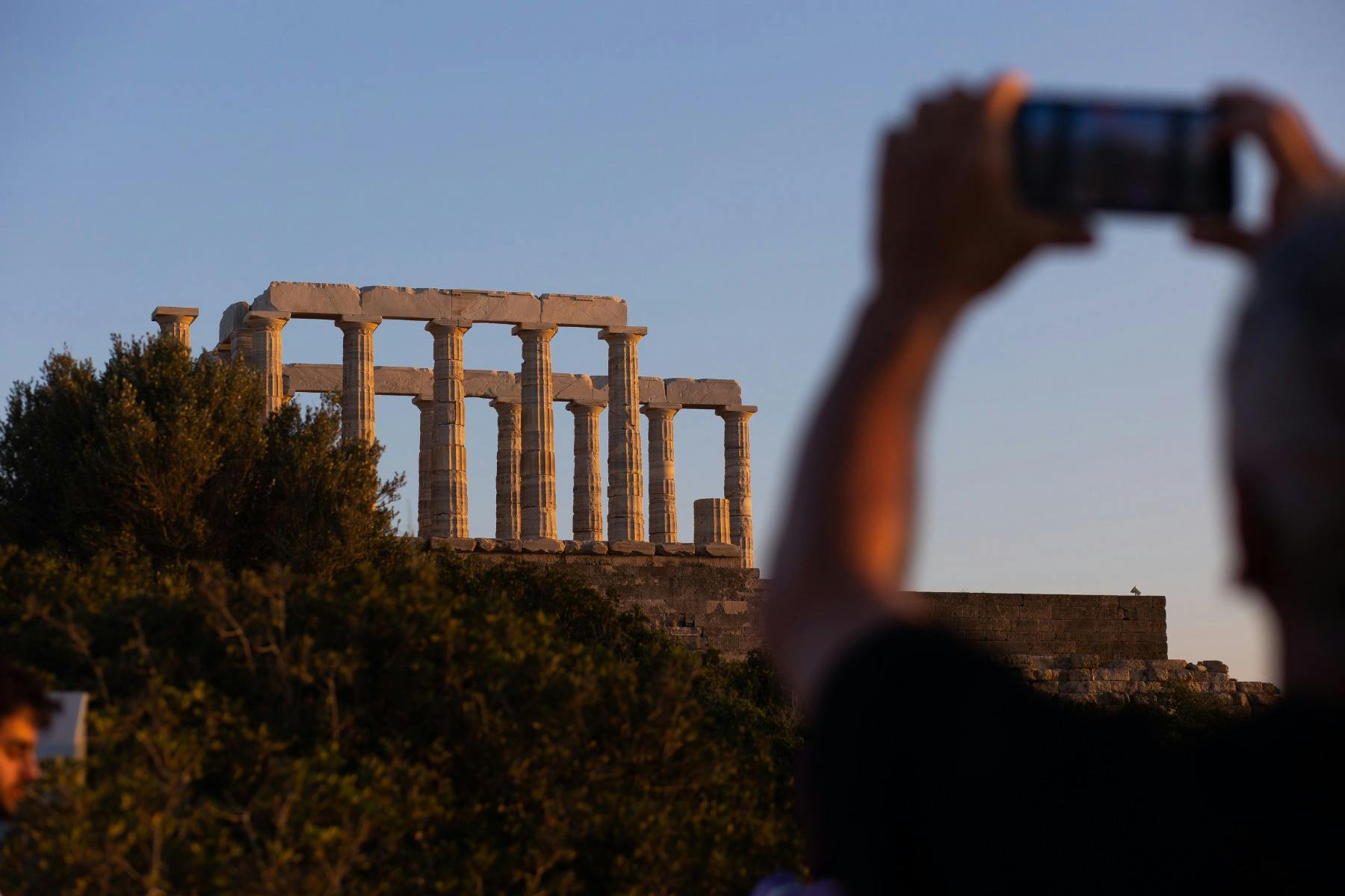A visitor takes a photo of the Temple of Poseidon bathed in soft golden sunset light.