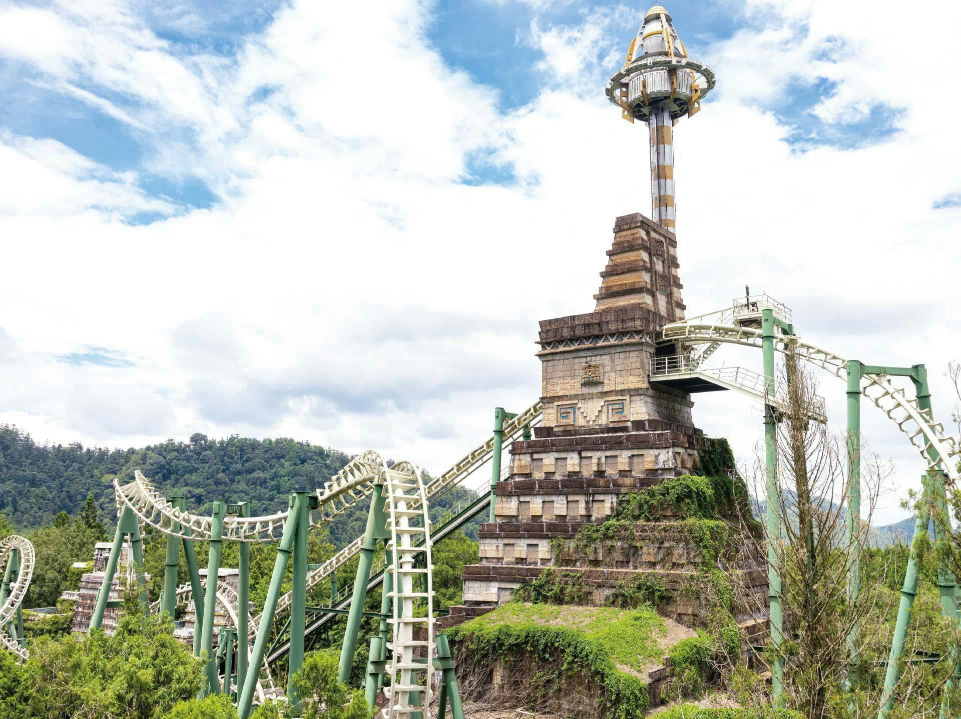 A roller coaster track winding around a tower with a spire, surrounded by greenery under a cloudy sky.