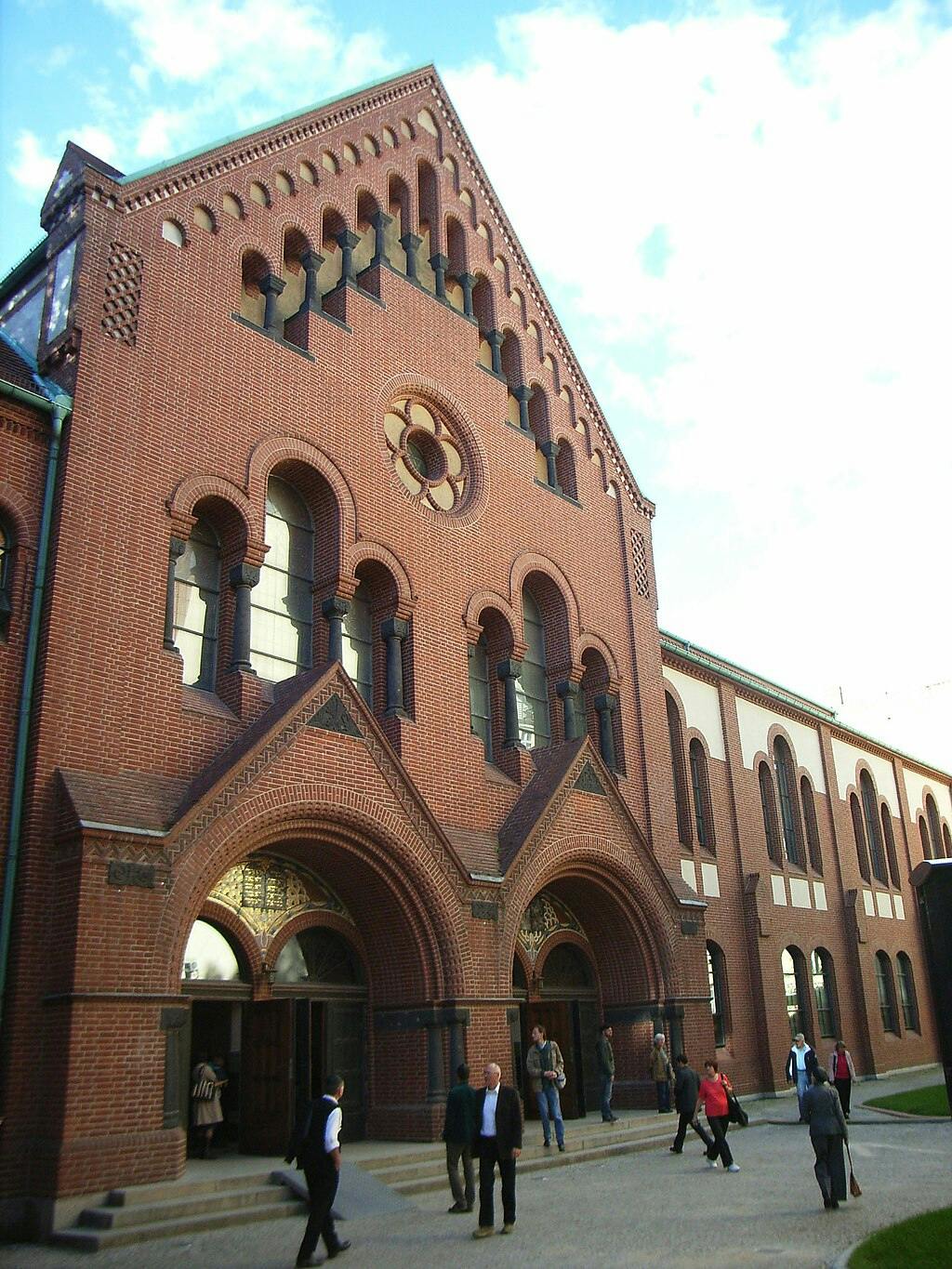 People walking in front of a large, ornate, red brick building with arched doorways and Gothic architectural details.