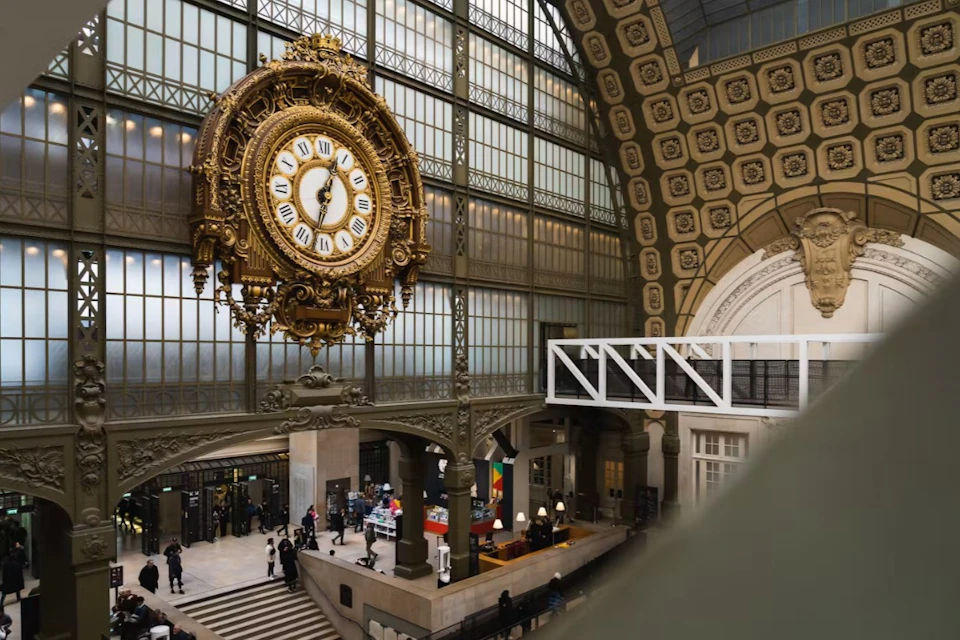 An ornate clock hangs in a spacious interior with high windows, decorative architecture, and people walking below.