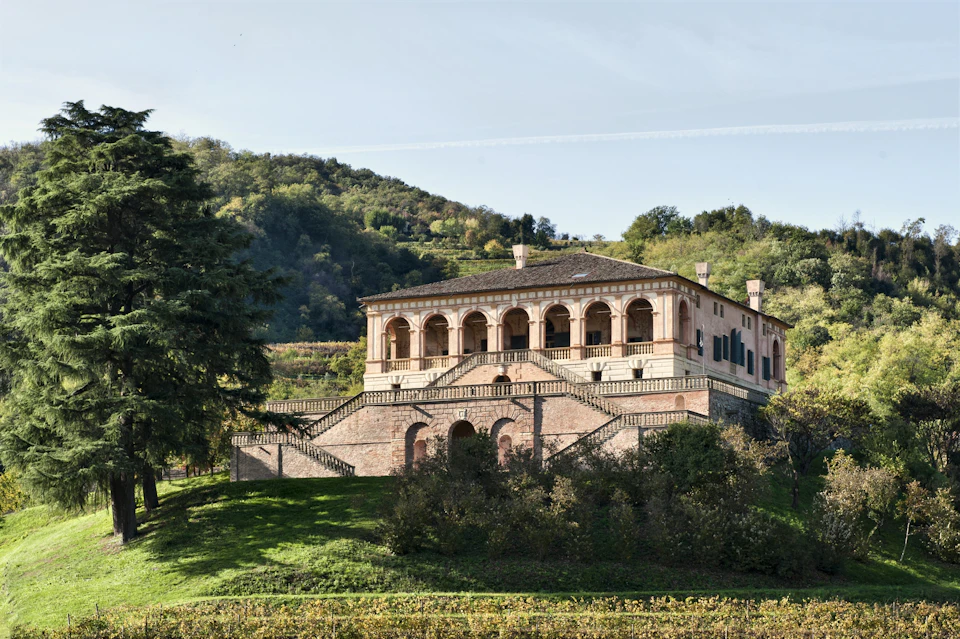 A large, historic villa with arches and multiple staircases on a green hillside, surrounded by trees and vegetation.