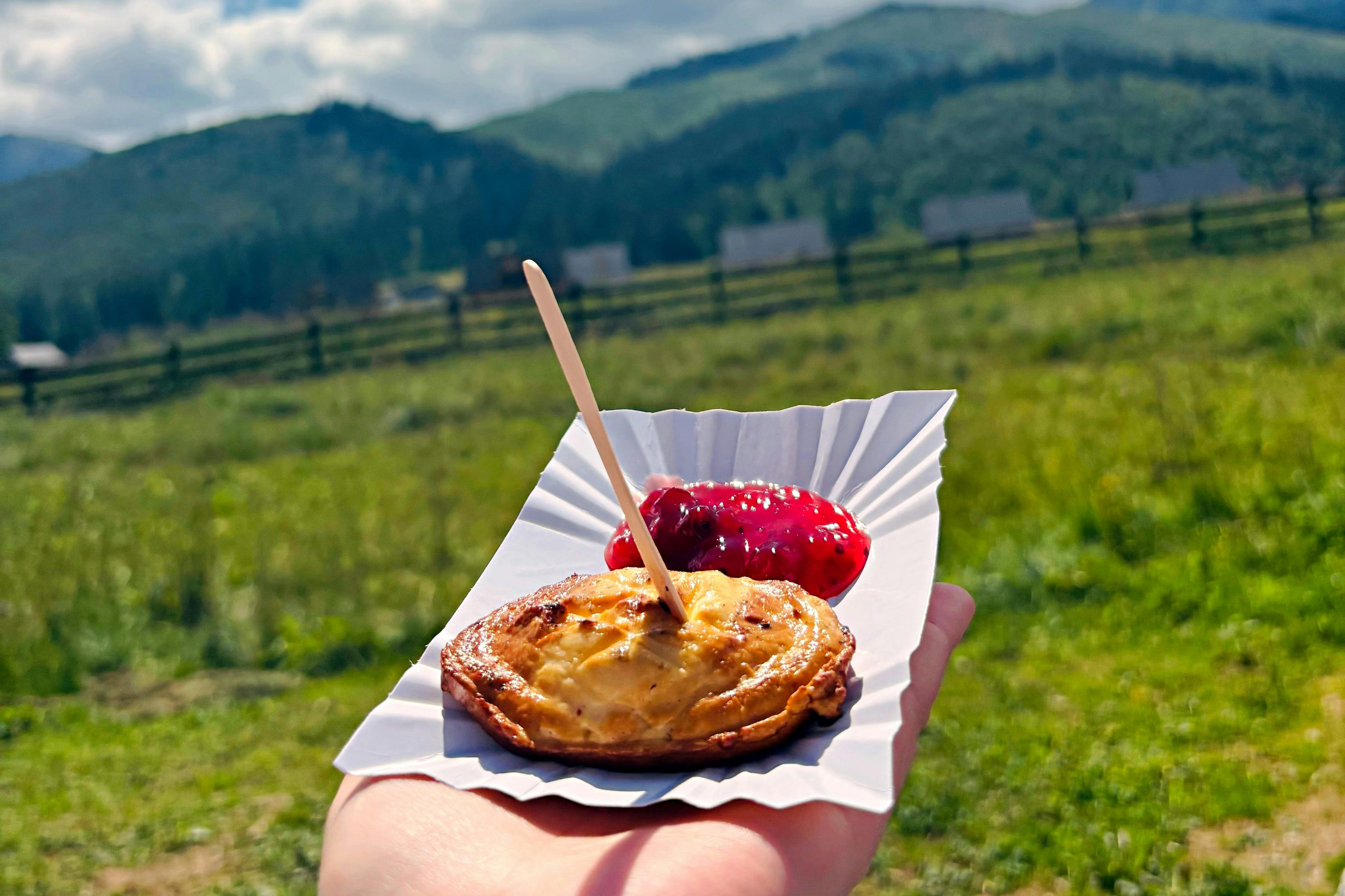 緑の山々と野原を背景に、木の棒が挿入された小さなペストリーと赤いソースが入ったトレイを手が持っています。