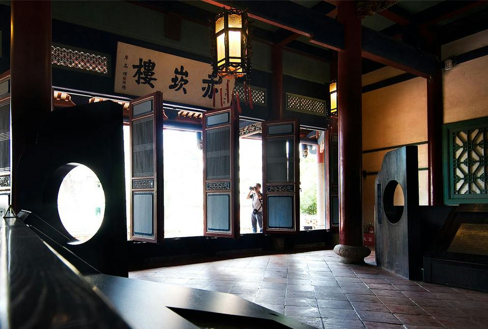 A traditional interior with open wooden doors, hanging lanterns, and a person standing outside in the sunlight.