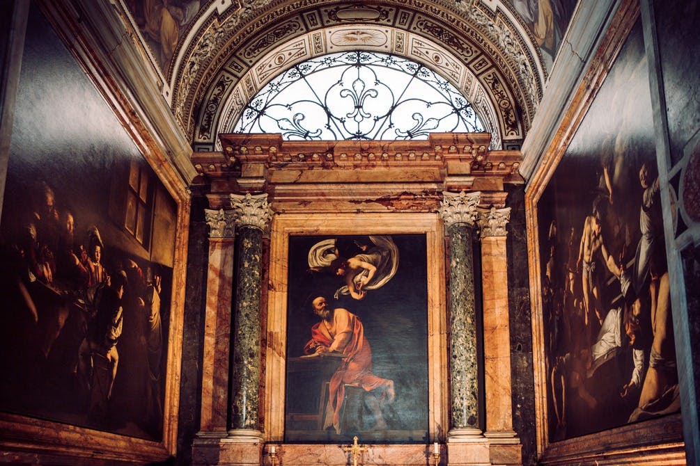 Interno di cappella ornato con un grande dipinto di una scena religiosa affiancato da colonne di marmo e intricati dettagli del soffitto.