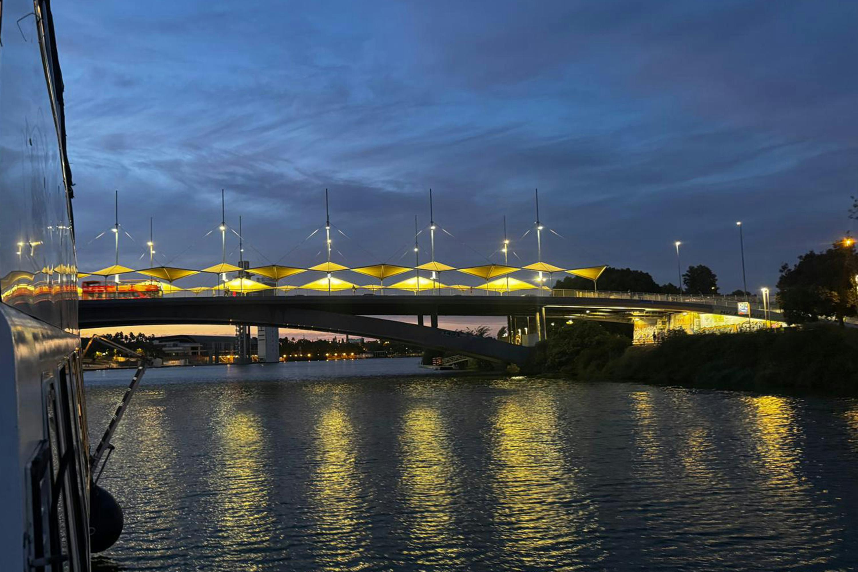Most o oświetlonych żółtych budowlach rozpościera się nad rzeką o zmierzchu, z zabudowaniami i drzewami w tle.