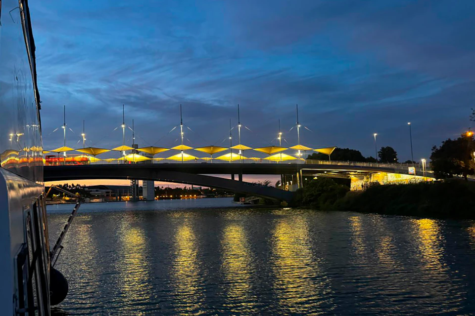 A bridge with illuminated yellow structures spans over a river at dusk, with buildings and trees in the background.
