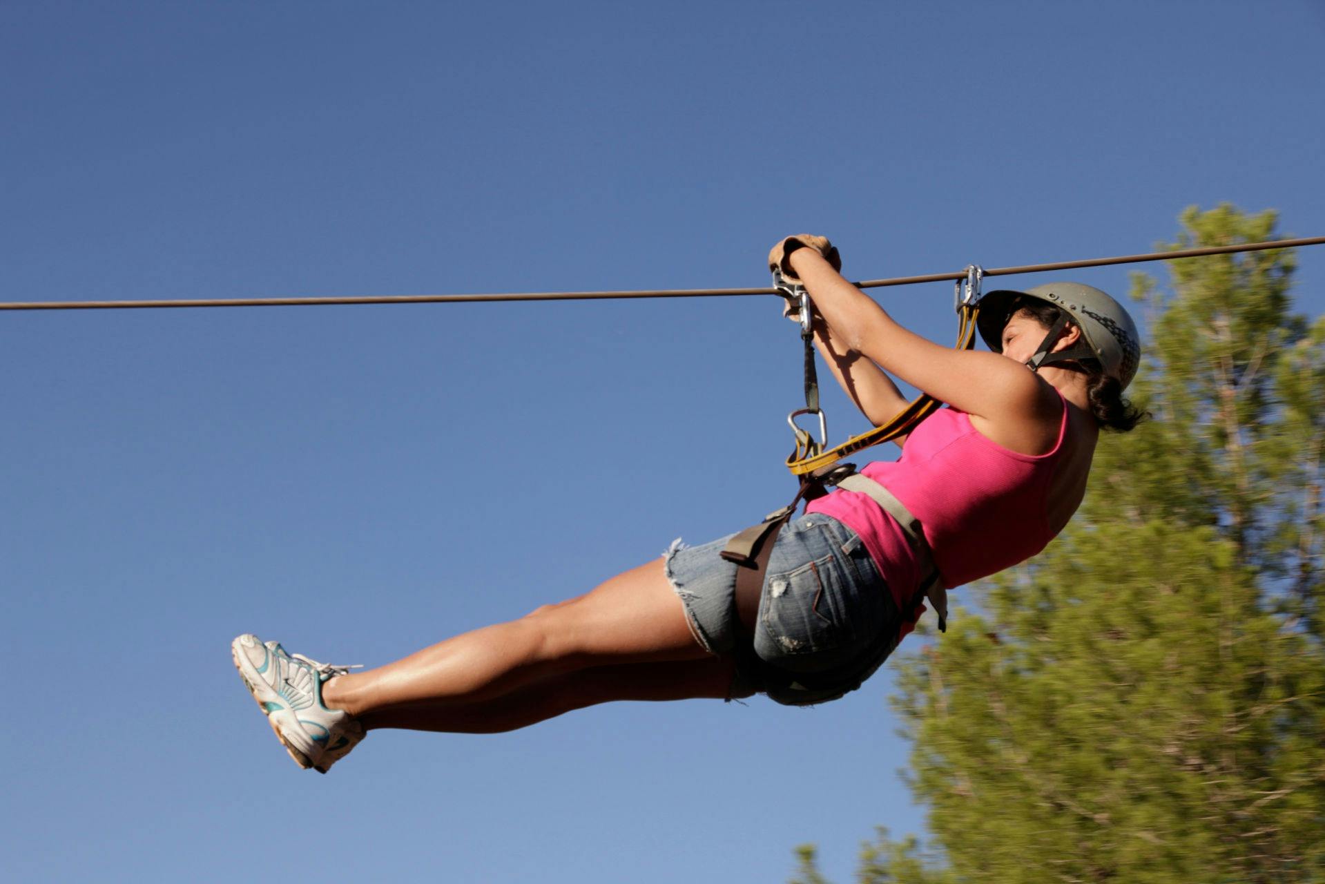 Person in a pink tank top and helmet gliding on a zipline against a clear blue sky, with some greenery visible.