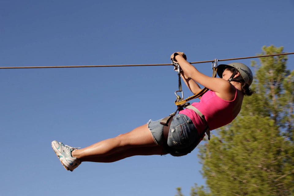 Person in a pink tank top and helmet gliding on a zipline against a clear blue sky, with some greenery visible.