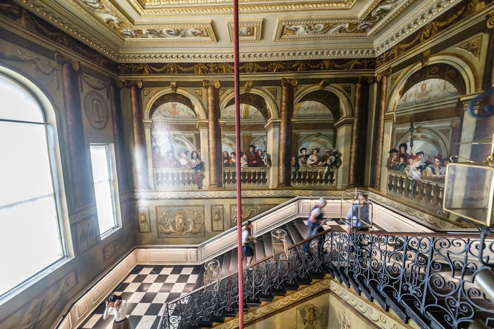People walking up a grand staircase with ornate decorations, murals, and black-and-white checkered flooring.