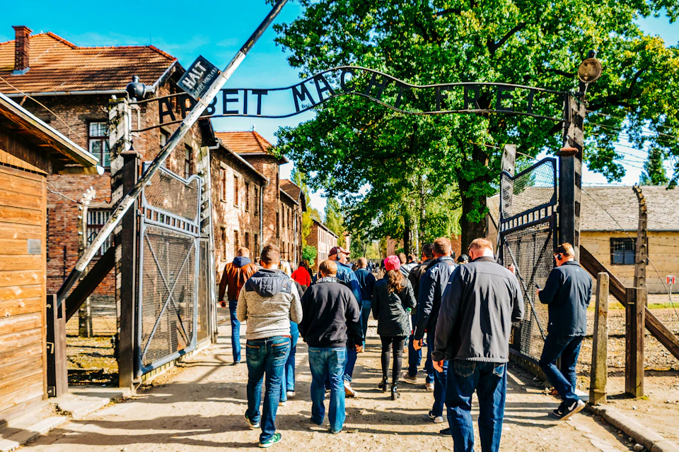Visitors walking under the "Arbeit Macht Frei" sign at the entrance gate of Auschwitz concentration camp, with trees and buildings nearby.