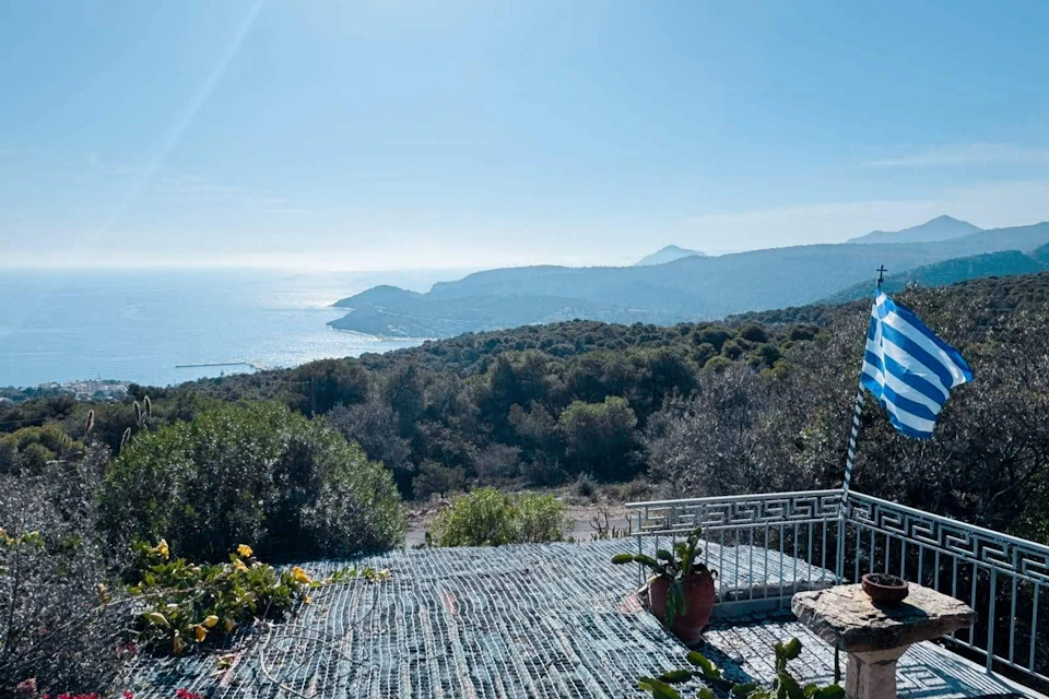 A scenic coastal view featuring hills, a calm sea, and a terrace with potted plants under a clear blue sky.