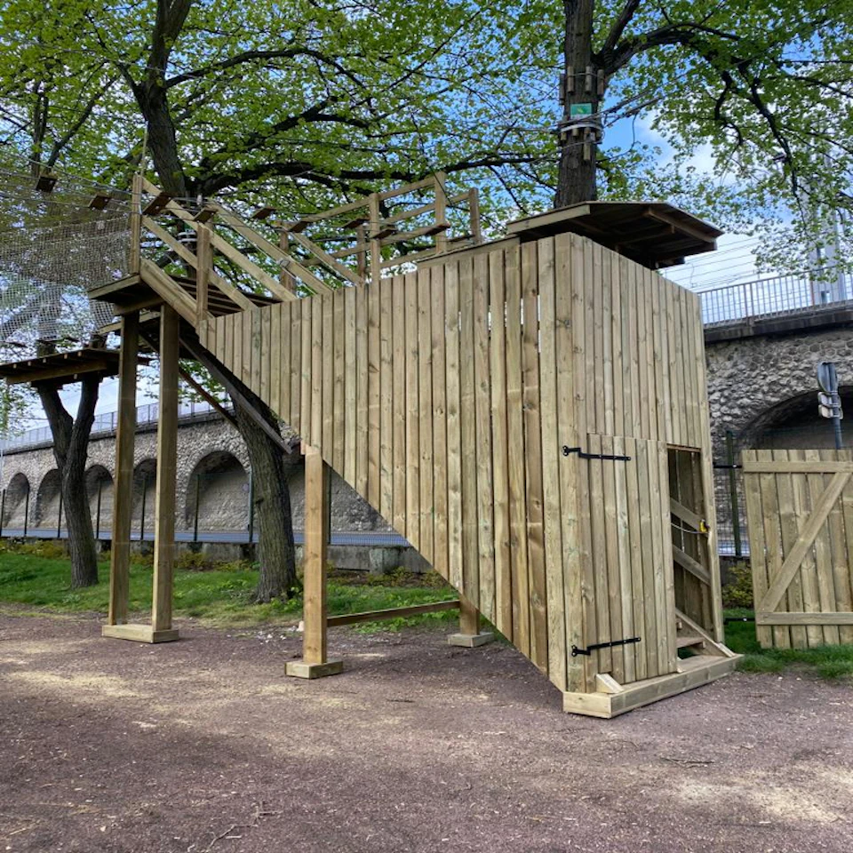 Wooden platform structure with stairs and a doorway, supported by posts. Located outdoors near trees and a stone wall with arches.
