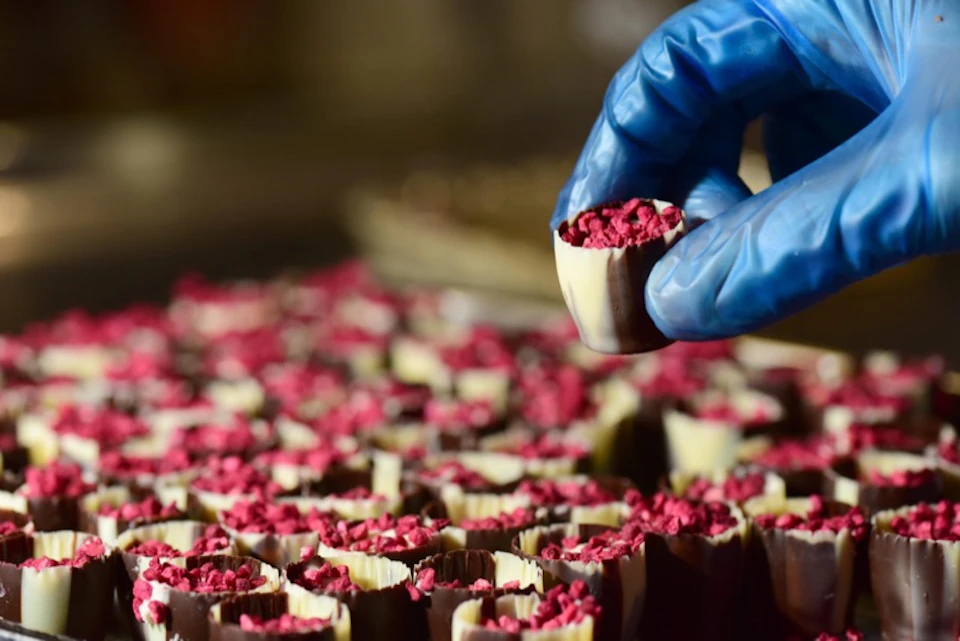 A gloved hand holding a small dessert filled with red toppings, with many similar desserts arranged in the background.