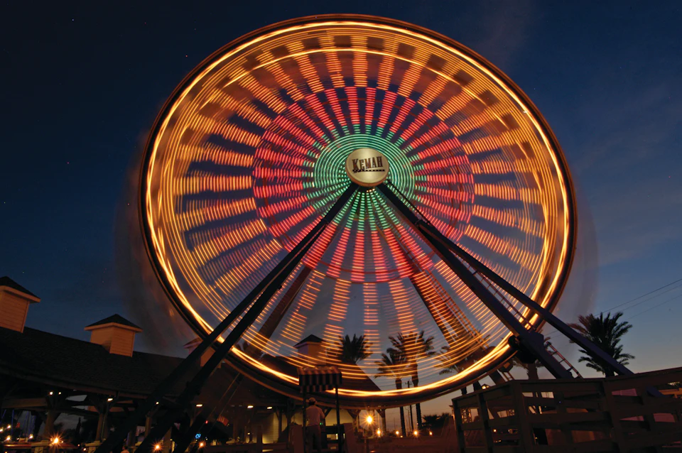 A Ferris wheel illuminated with colorful lights spins against a dark evening sky, with palm trees and buildings in the background.
