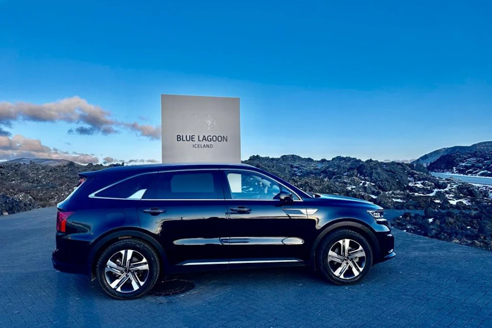 A sleek black SUV is parked in front of a sign that reads "Blue Lagoon Iceland," with a clear blue sky in the background.