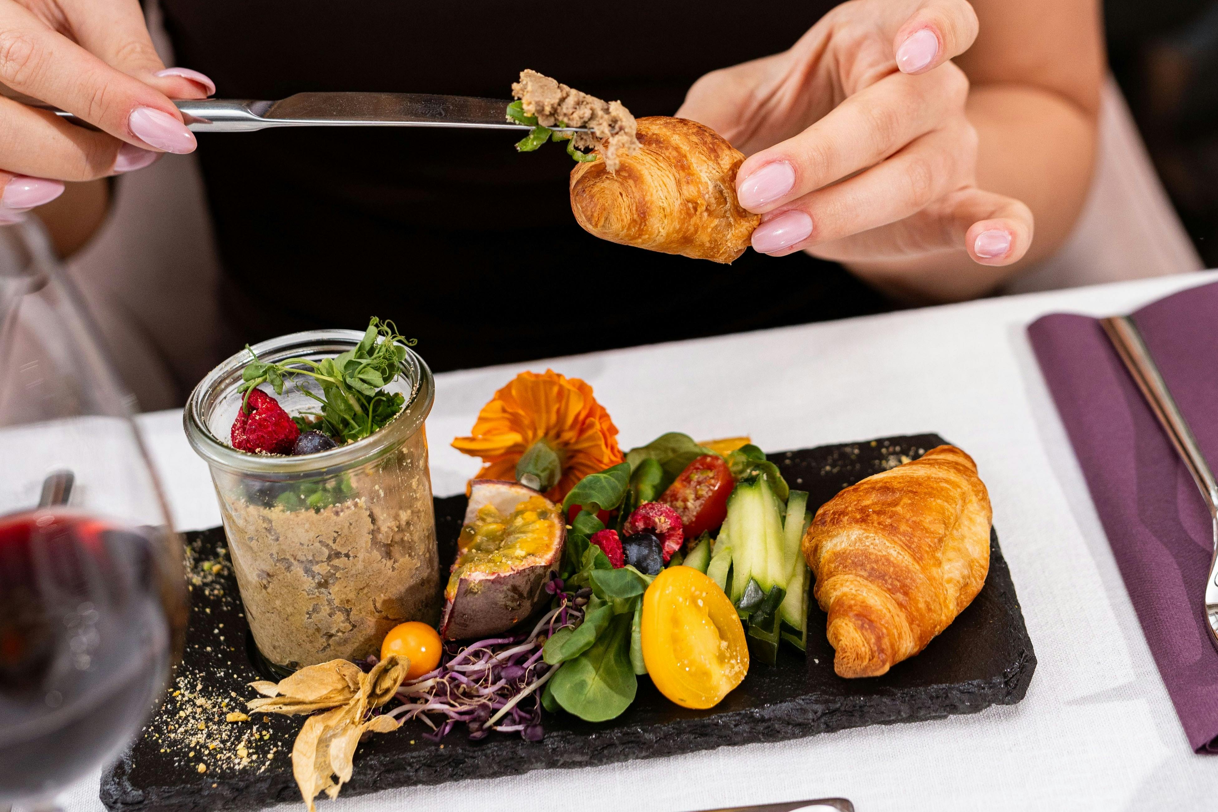 Una persona estenent una pasta sobre un petit croissant en un plat amb un colorit arranjament de fruites, verdures i flors.