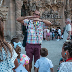 A tour guide in a plaid shirt gestures while speaking to a group of children outside an intricately detailed stone building.