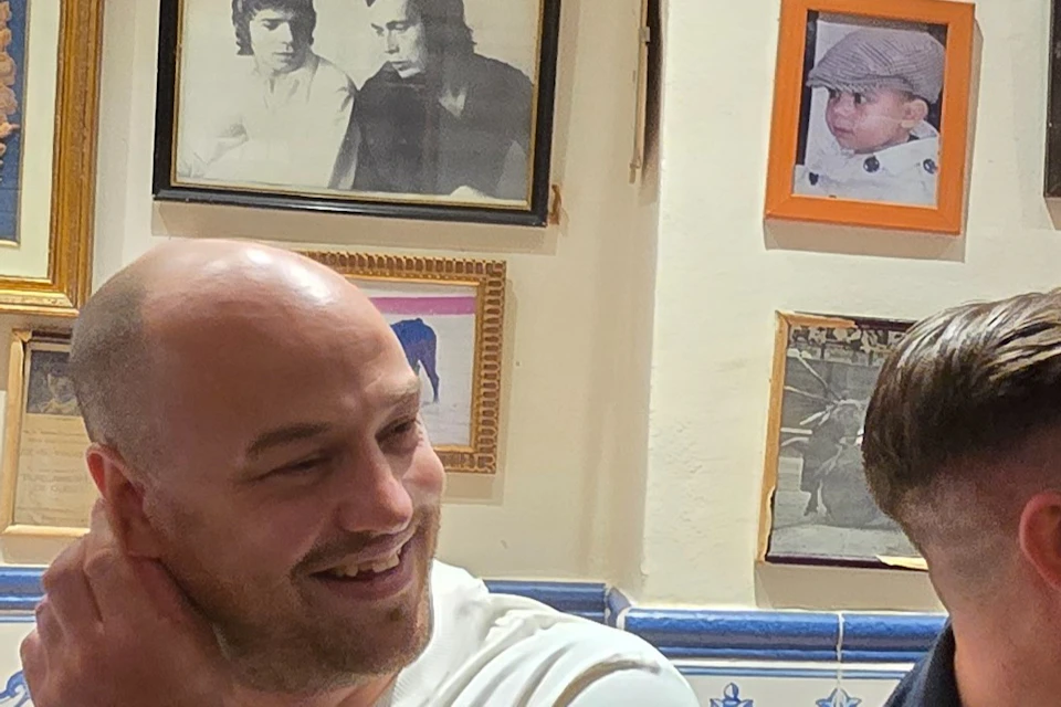 Four men sitting in a room with framed photos on the wall, laughing and chatting. One holds a glass of wine.