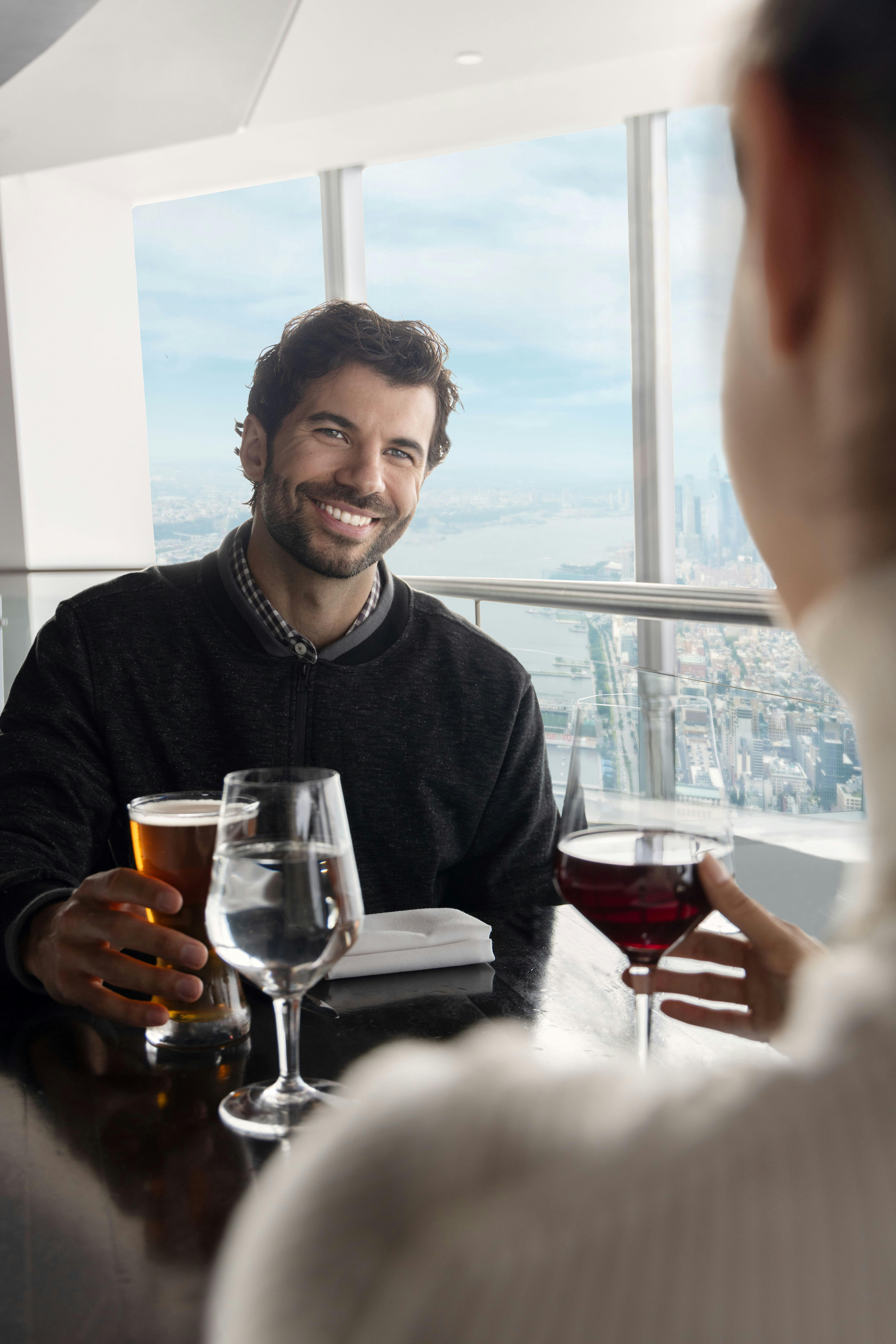 赤ワインを飲んでいる人の向かい側にビールを持っている男性が微笑んでいます。 シティビューの窓のそばに座っています。
