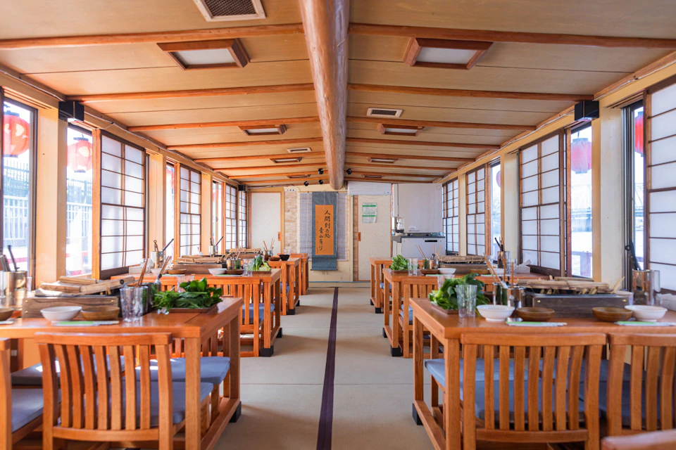 A spacious Japanese-style dining room with wooden tables and chairs, shoji screens, and neatly set dining utensils and plants.