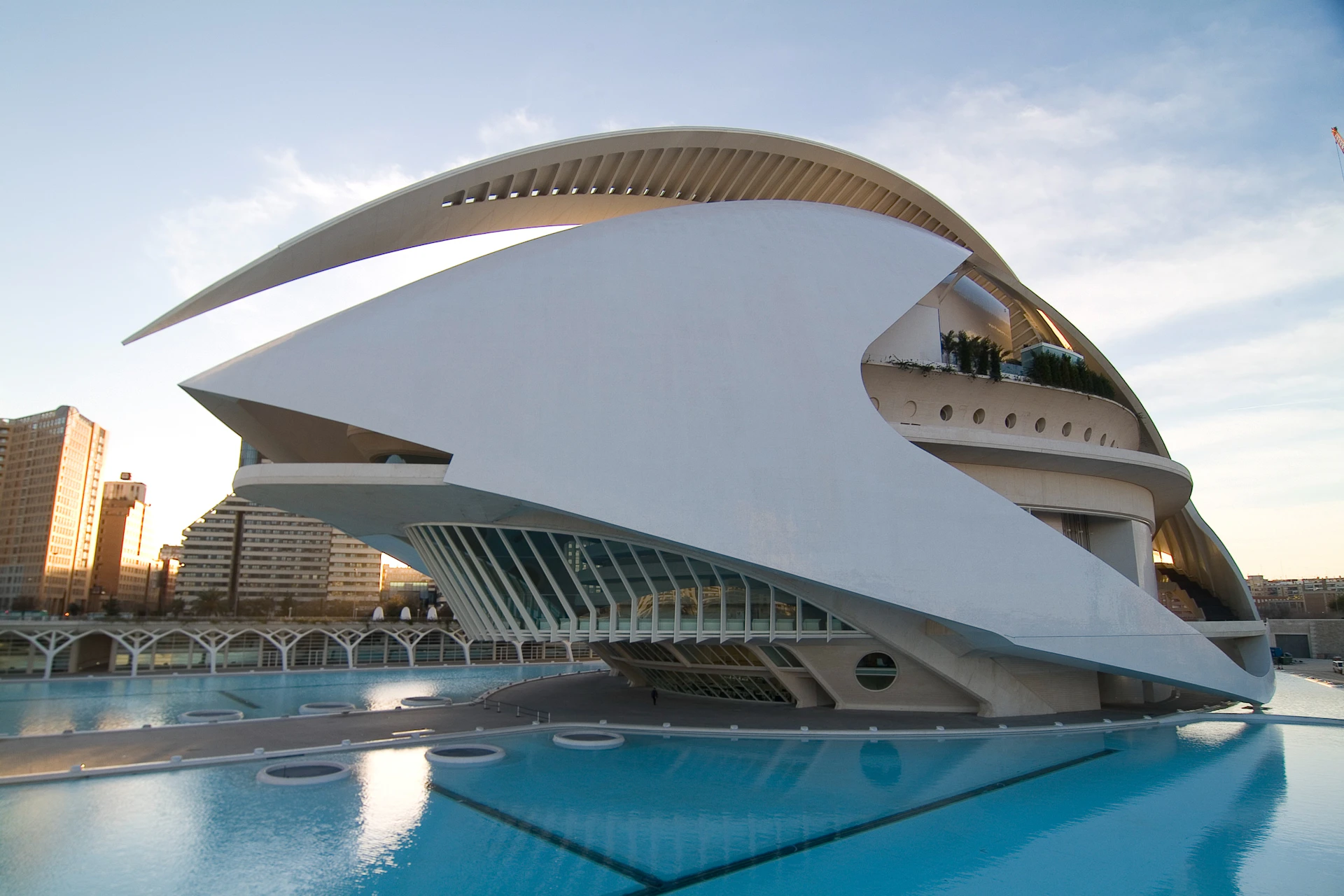 Futuristic white building with curved architecture over a reflective pool, set against a clear sky.