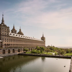 En stor historisk byggnad med kupoler och spiror har utsikt över en reflekterande pool och välskötta grunder under en klar himmel.